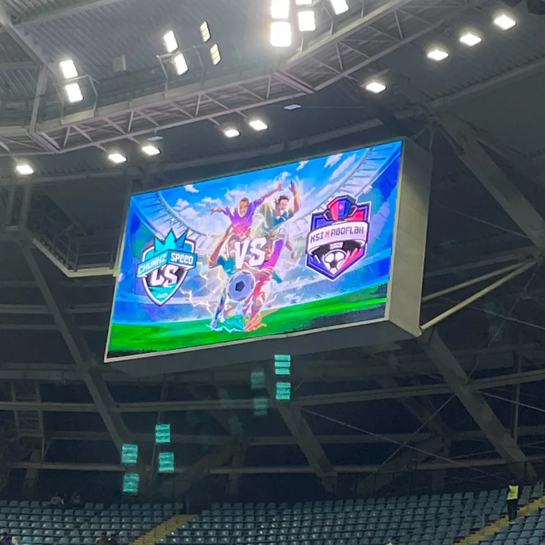 A large stadium screen displays colorful team logos and animated characters for a football match between "Chennai Speed" and "East Adolph," highlighting nation branding as bright lights illuminate the scoreboard.