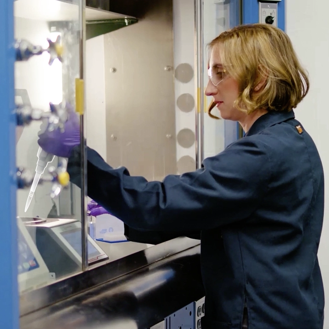 A person in a lab coat works inside a laboratory fume hood, using a pipette. They are wearing safety goggles and purple gloves, focusing on an experiment or procedure. Various equipment and controls are visible around them.