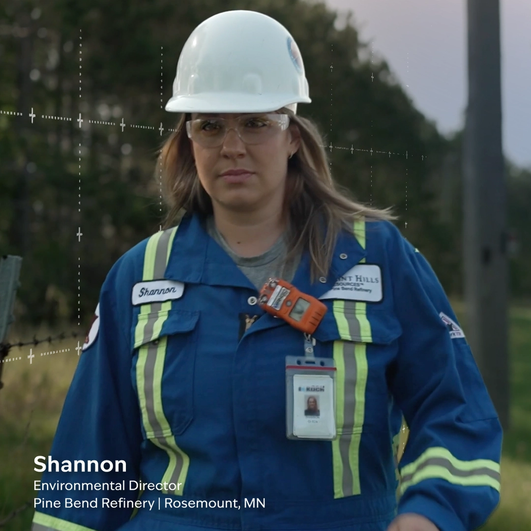 A person in protective gear stands outdoors. They wear a white hard hat, safety glasses, and a blue work uniform with a visible ID badge. Text reads: "Shannon, Environmental Director, Pine Bend Refinery, Rosemount, MN.