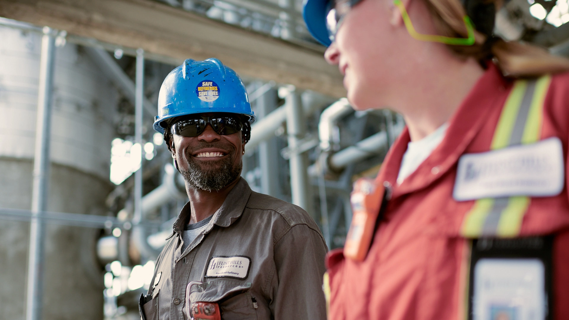 Two industrial workers in helmets and safety gear are conversing. One, in a blue helmet, smiles at the other, who wears a red helmet. They are in an outdoor industrial setting with blurred piping in the background.