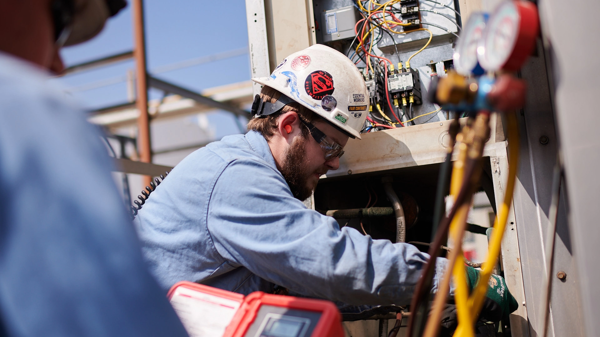 A technician wearing a hard hat and safety goggles works on electrical components inside an open panel. They are surrounded by tools and wires, focusing intently. The background shows industrial equipment and a clear blue sky.