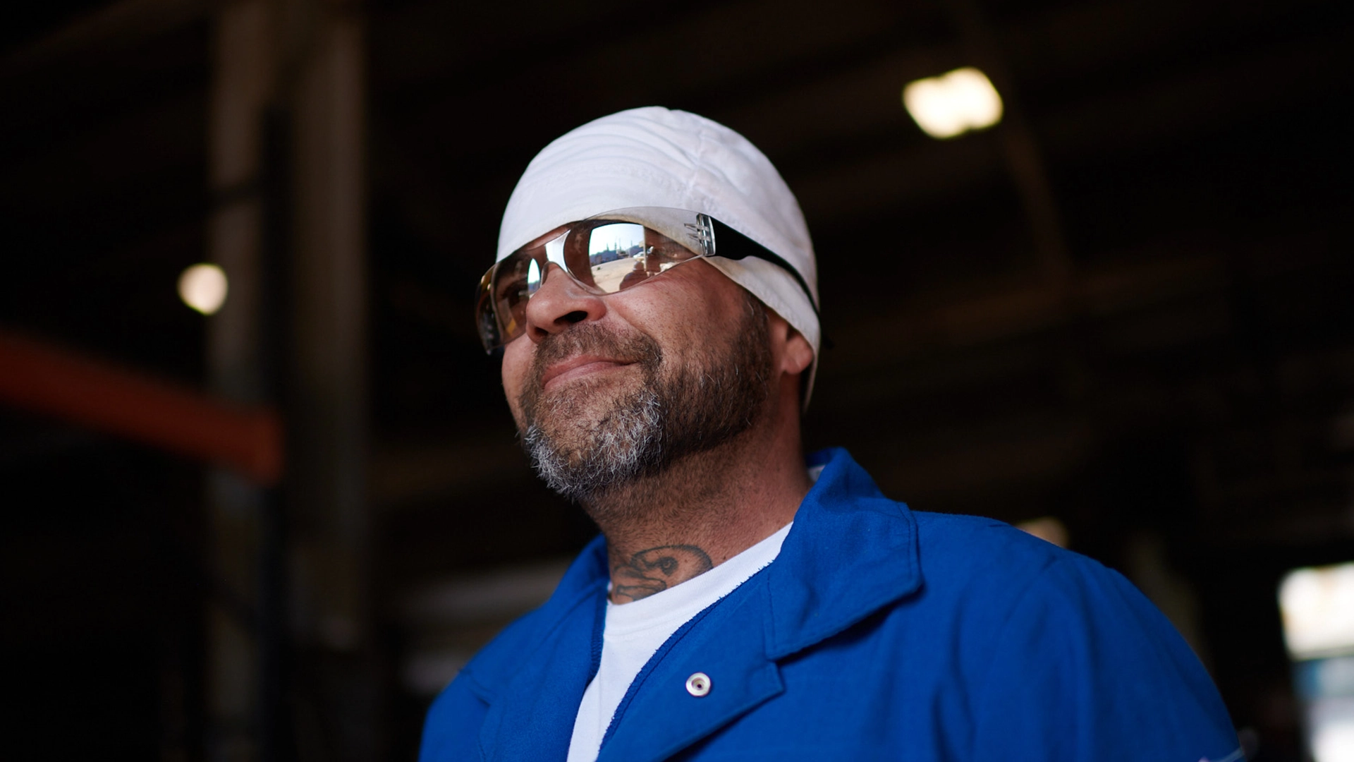 A man with a beard wearing a white bandana, sunglasses, and a blue work shirt stands indoors, smiling slightly. The background is dark and out of focus.