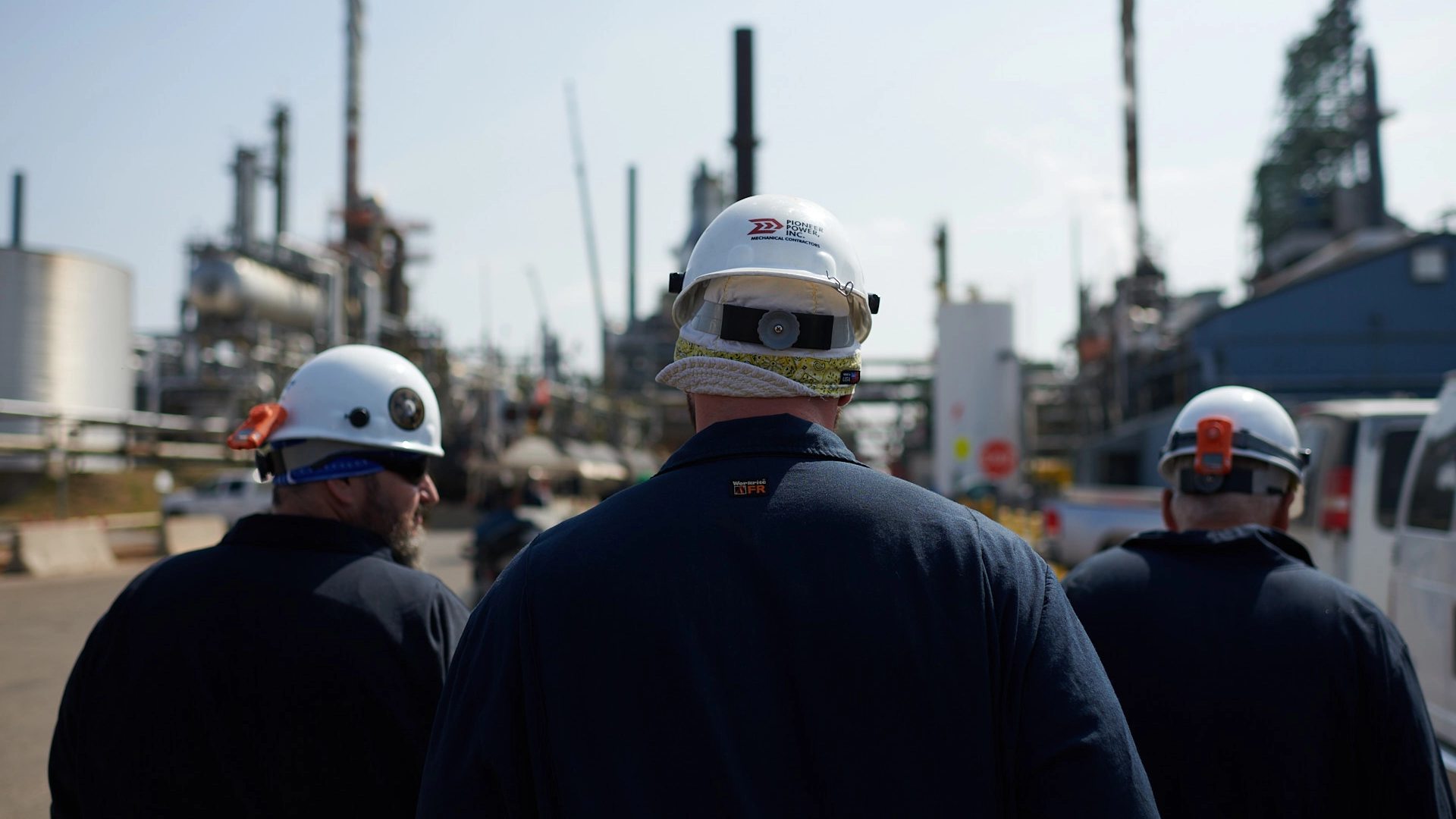 Workers in safety helmets walk through an industrial site with pipes and structures in the background. They appear to be inspecting or discussing the area. The sky is clear and the setting is outdoors.