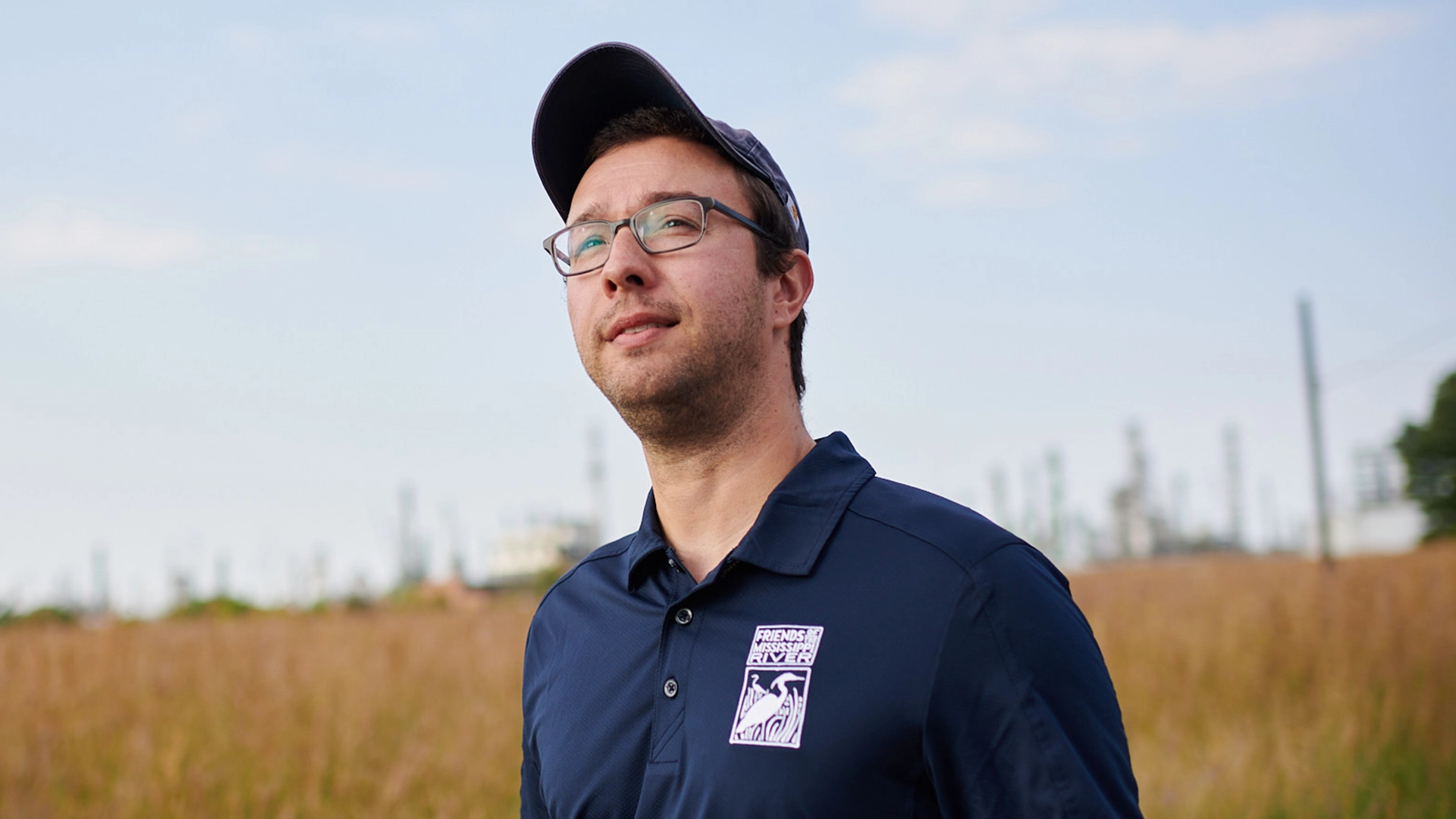 A person wearing glasses and a navy cap stands in a field, looking into the distance. They are wearing a navy shirt with a logo. The background features tall grass and an industrial area under a blue sky.