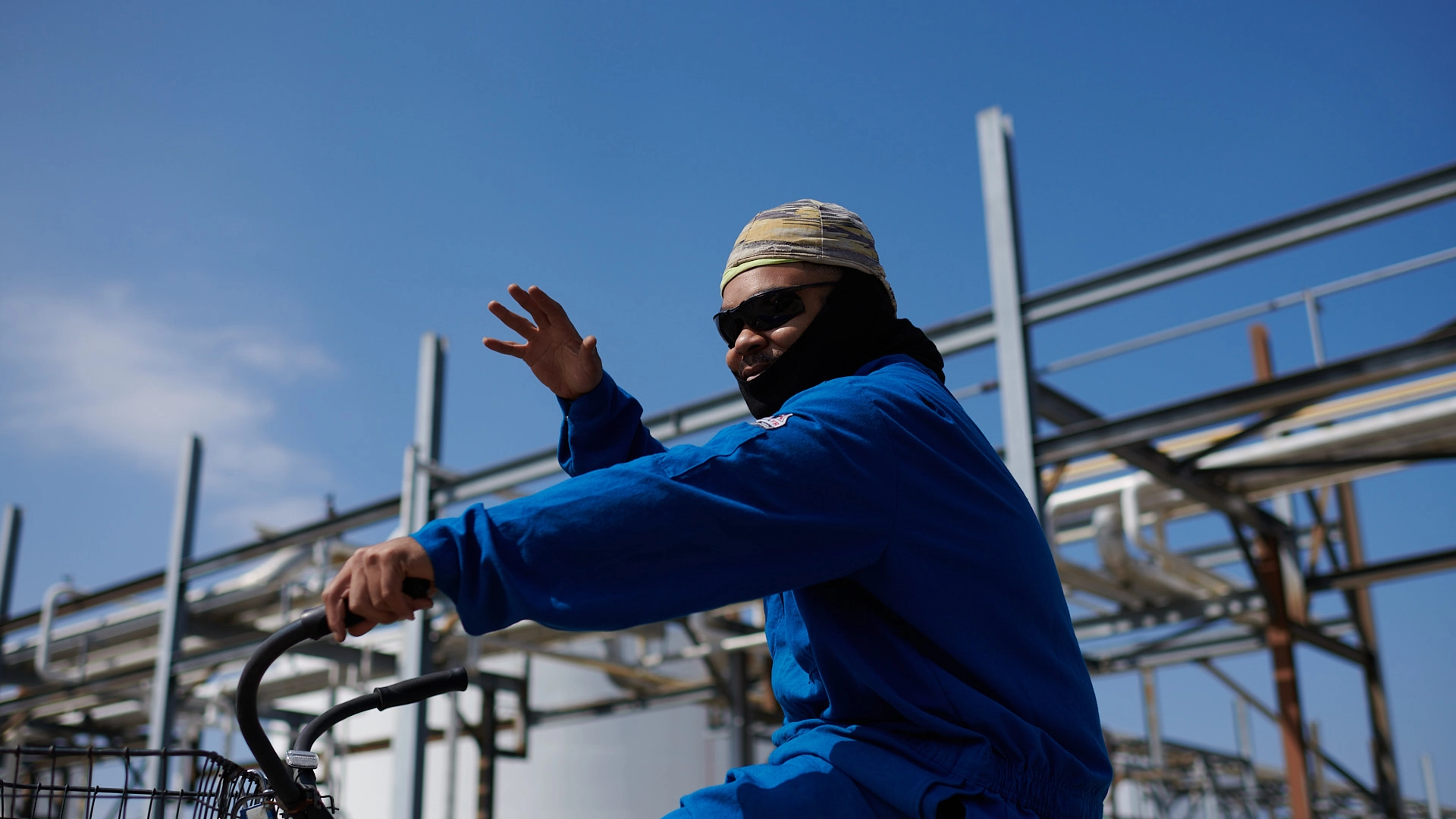 A person wearing a blue jumpsuit, headscarf, and sunglasses rides a bicycle, waving with one hand. Background features a bright blue sky and industrial metal structures.