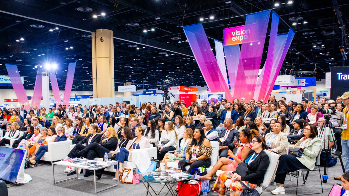 A large audience sits and listens to a speaker at an indoor expo event, surrounded by colorful banners and exhibition booths with "Vision Expo" signage, highlighting the importance of health advocacy in public policy communications.