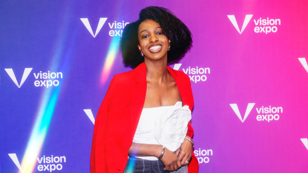 A person smiling in front of a colorful Vision Expo backdrop, wearing a red blazer draped over their shoulders, a white top with a floral detail, and blue jeans—embodying the spirit of health advocacy.