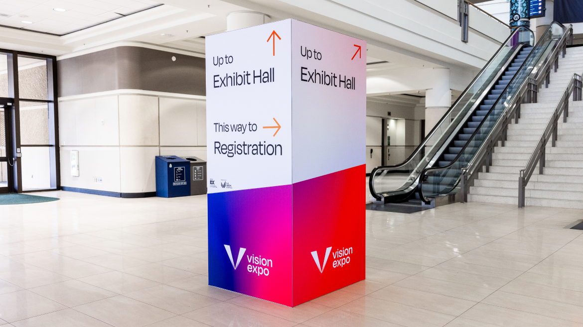 A large sign in a convention center lobby directs visitors to the Exhibit Hall upstairs and to Registration; promoting Vision Expo, it highlights effective communication strategy near an escalator and a staircase.