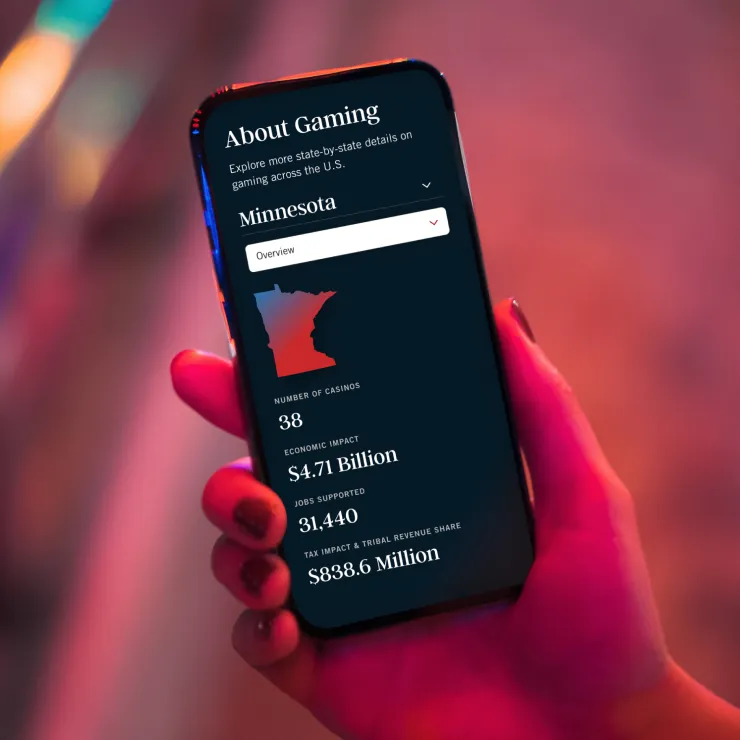 A hand holds a smartphone displaying gaming statistics for Minnesota, including 38 casinos, $4.71 billion economic impact, 31,440 jobs supported, and $838.6 million in tax and tribal revenue.