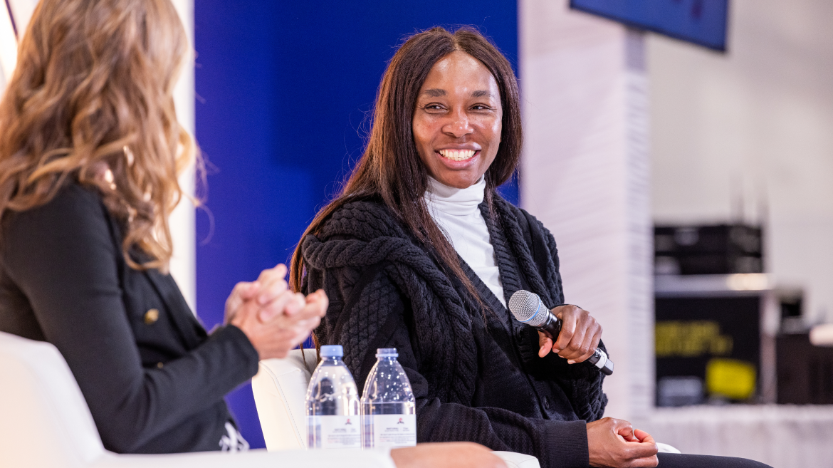 Two women sit and talk on stage. One smiles while holding a microphone, with two water bottles on the table before them. The blue and white background suggests a formal event focused on health communication or a panel hosted by PhRMA.