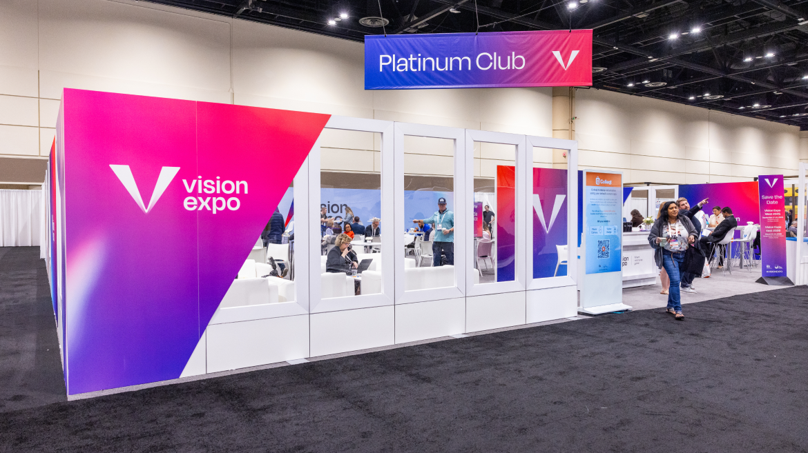 A bright exhibition booth with "Vision Expo" and "Platinum Club" signs, featuring glass walls and people discussing communication strategy inside and around the booth at a convention center.