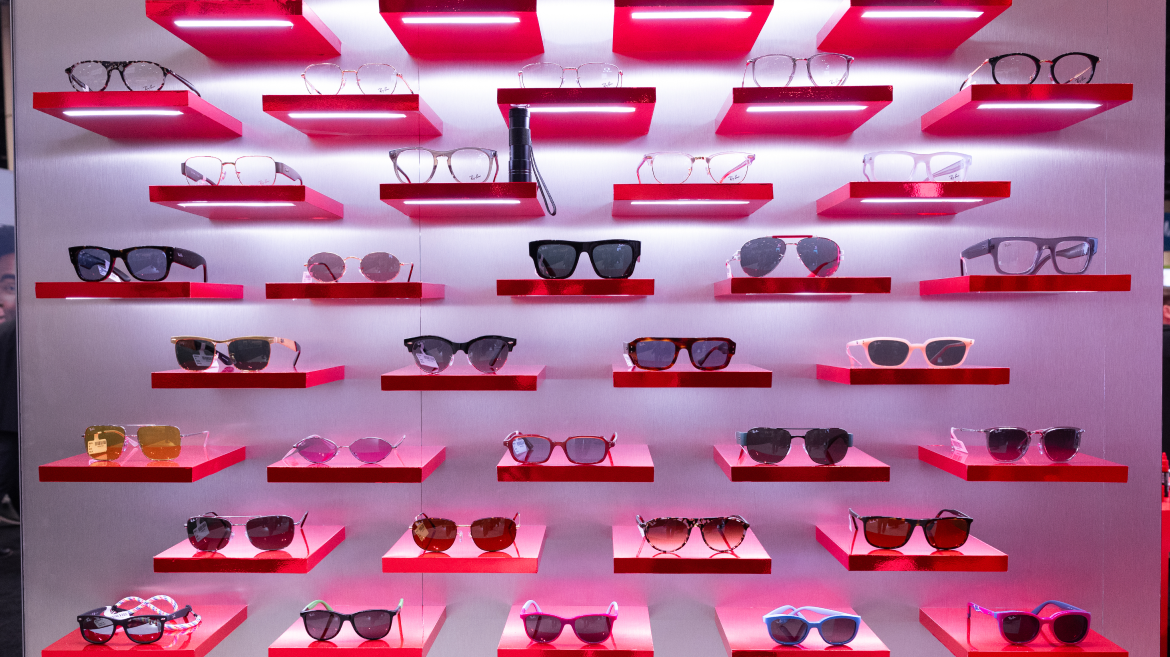 A display of various sunglasses and eyeglasses arranged on red, backlit shelves against a metallic wall, with rows of different frame styles and lens colors showcased to highlight the importance of health communication in eye care.