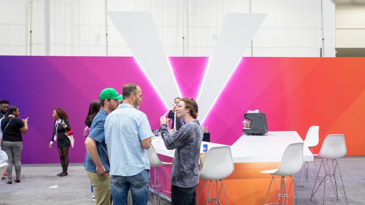 A group of people stand and talk near a modern bar-like counter with white stools, set against a vibrant, colorful backdrop featuring a large "V"—an inviting setting for discussions on health communication. Others mingle in the background of the event space.