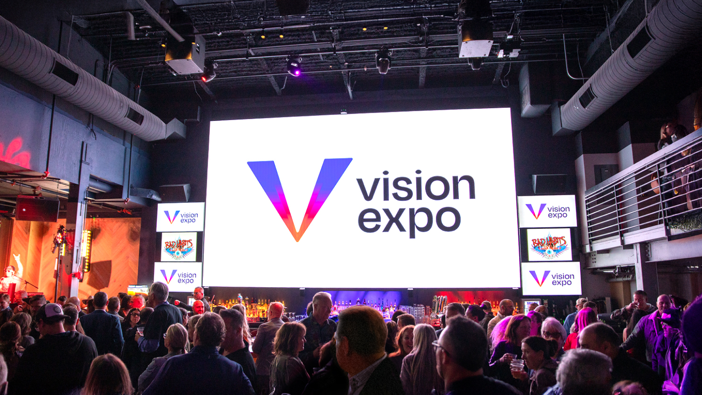 A large crowd gathers in front of a brightly lit screen displaying the “vision expo” logo inside a modern, indoor event space, highlighting the importance of health advocacy and effective communication strategy in eye care.