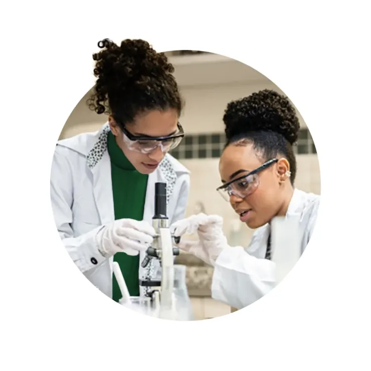 Two women wearing lab coats, safety goggles, and gloves look closely at a specimen through a microscope in a laboratory setting.