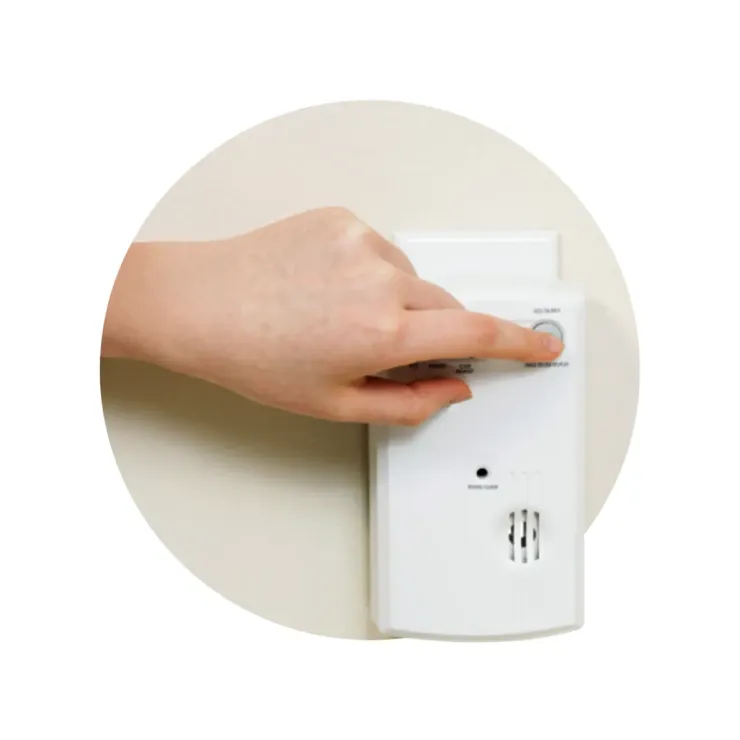 A hand pressing the test button on a wall-mounted carbon monoxide detector.