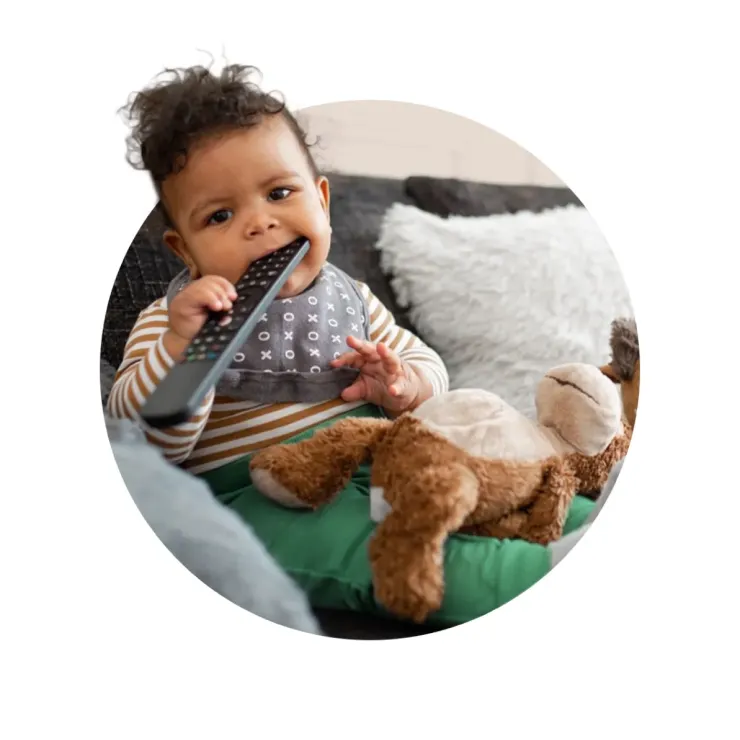 A baby sitting on a couch holds a stuffed animal and chews on a TV remote, wearing a striped shirt and a bib, with curly hair and a curious expression.
