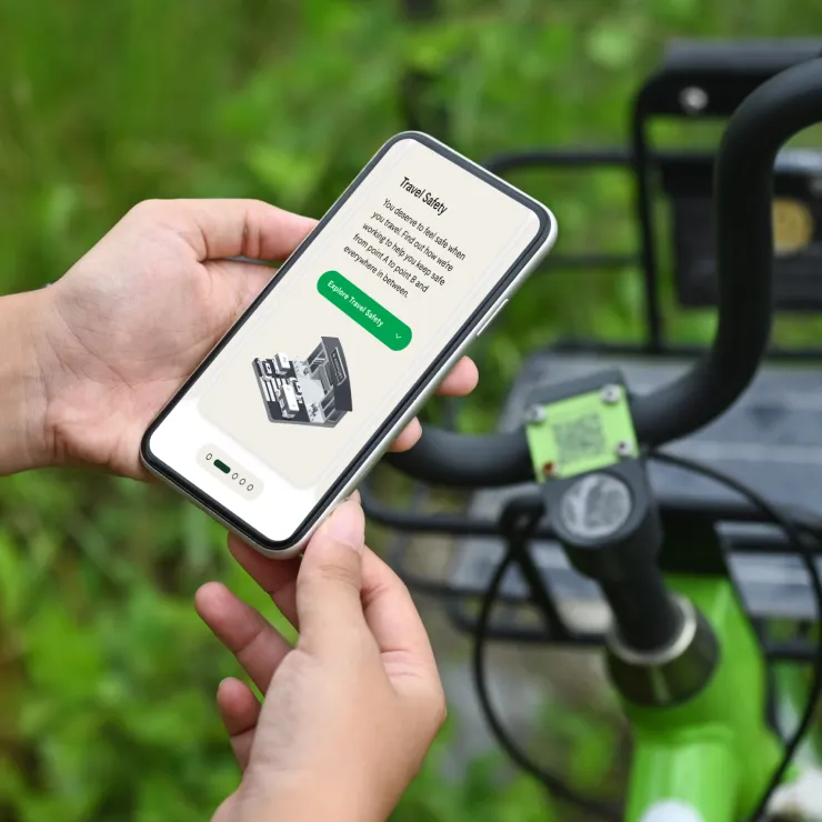 A person holds a smartphone displaying a travel safety app while standing next to the handlebars of a green bike outdoors, with greenery in the background.
