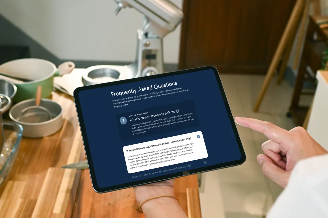 A person in a kitchen holds a tablet displaying a FAQ page about carbon monoxide poisoning, with text and answers shown on the screen.