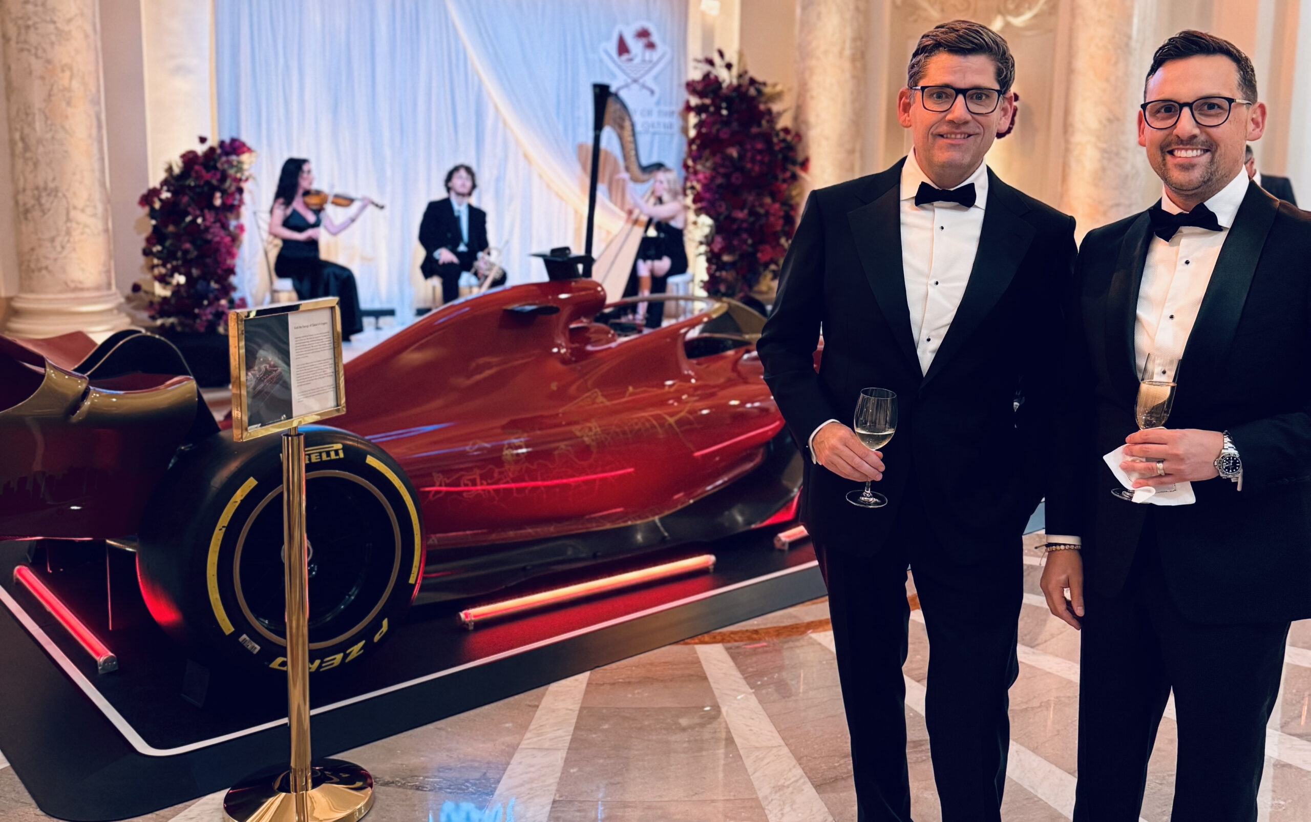 Two men in tuxedos hold drinks beside a red Formula 1 car display at an elegant indoor event. In the background, a string quartet performs, setting the stage for a creative public affairs strategy that begins with "Our Story.