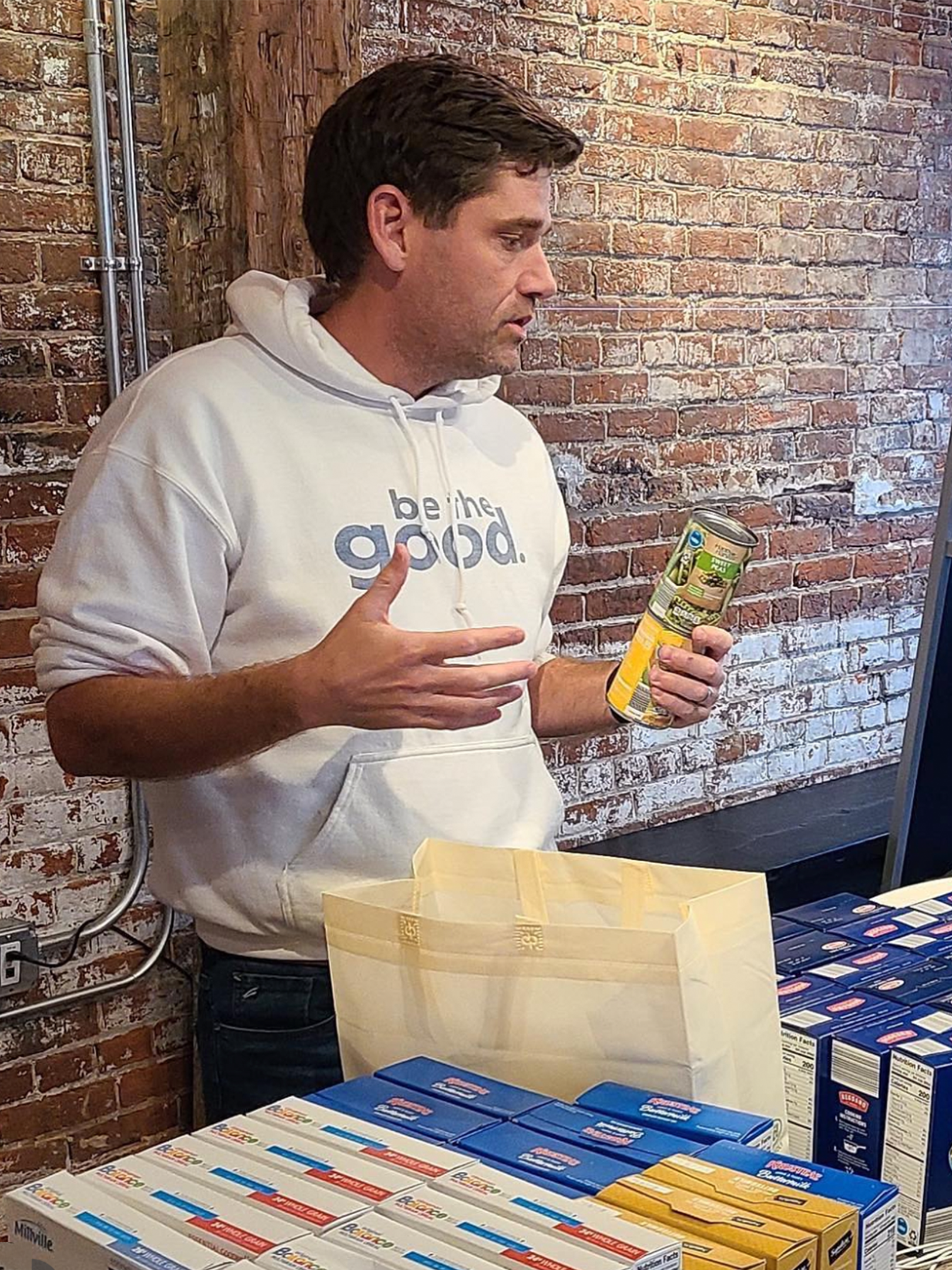 A man in a white "be the good." hoodie stands indoors by a table with food items, holding a canned good and gesturing with his other hand. Brick wall background, grocery boxes and a paper bag on the table.