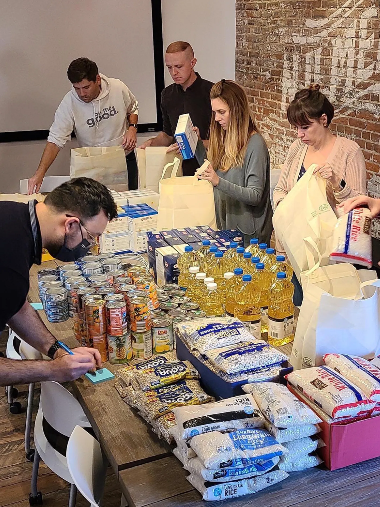 Five people stand around a table filled with canned goods, cooking oil, rice, and pasta, packing food items into tote bags as part of a volunteer activity in a room with brick walls.