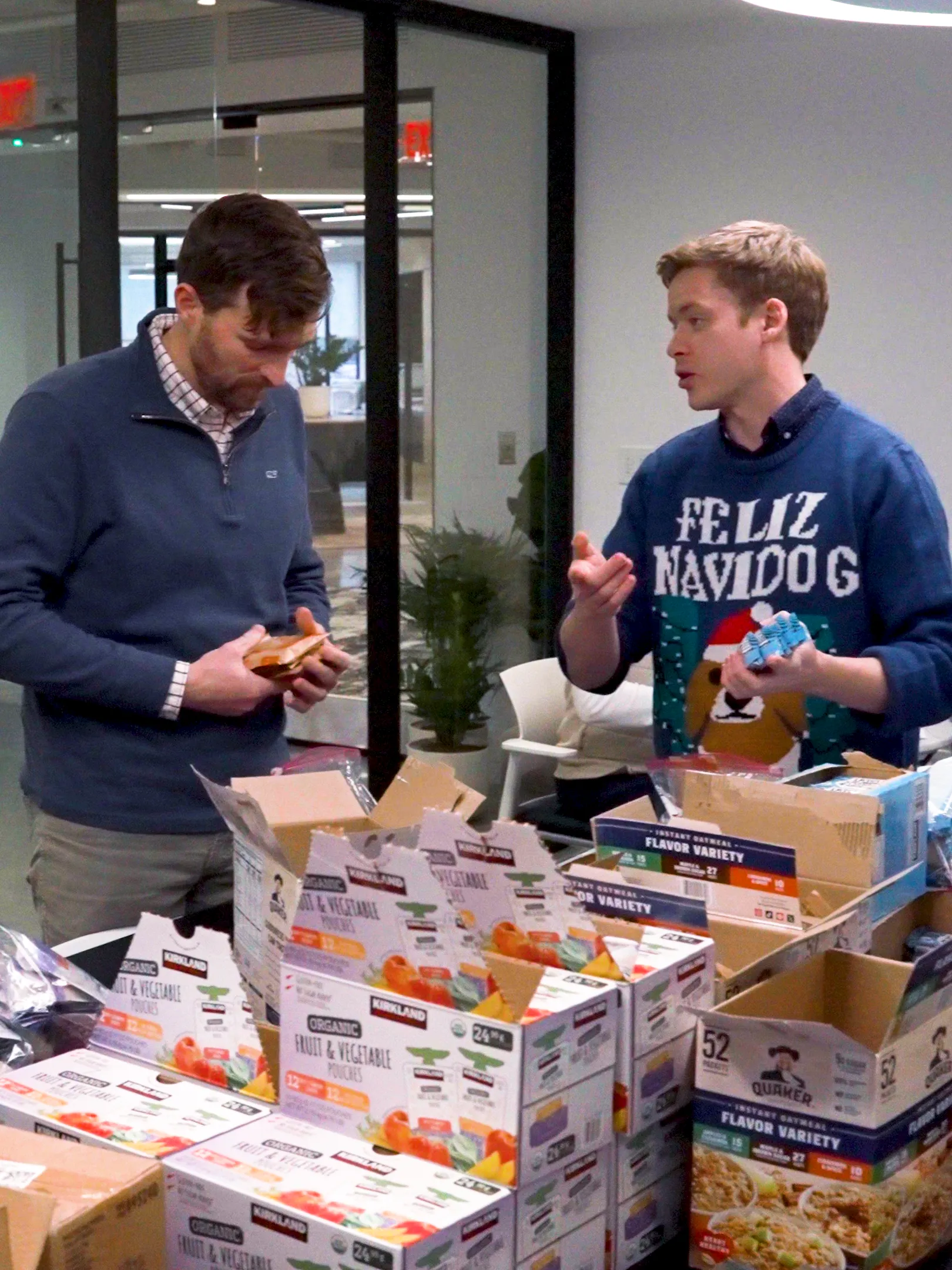 Two men stand at a table packing food into boxes. One man wears a blue sweater with "Feliz Navidog" and a dog graphic, while the other wears a plaid shirt. The table is covered with boxes of assorted food items.