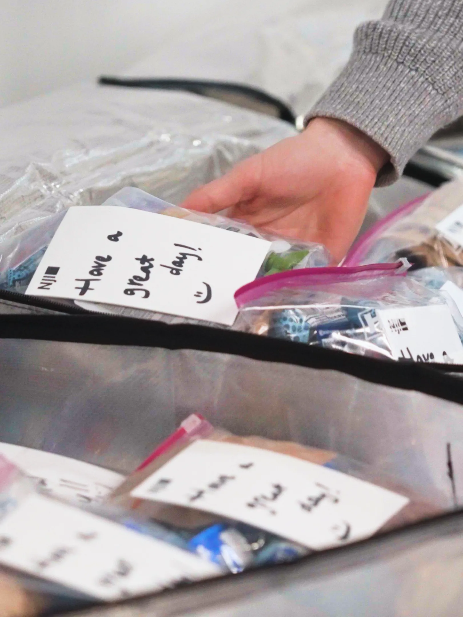 A hand places a ziplock bag containing snacks and a note that says "Have a great day! :)" into a larger bag filled with similar packages.