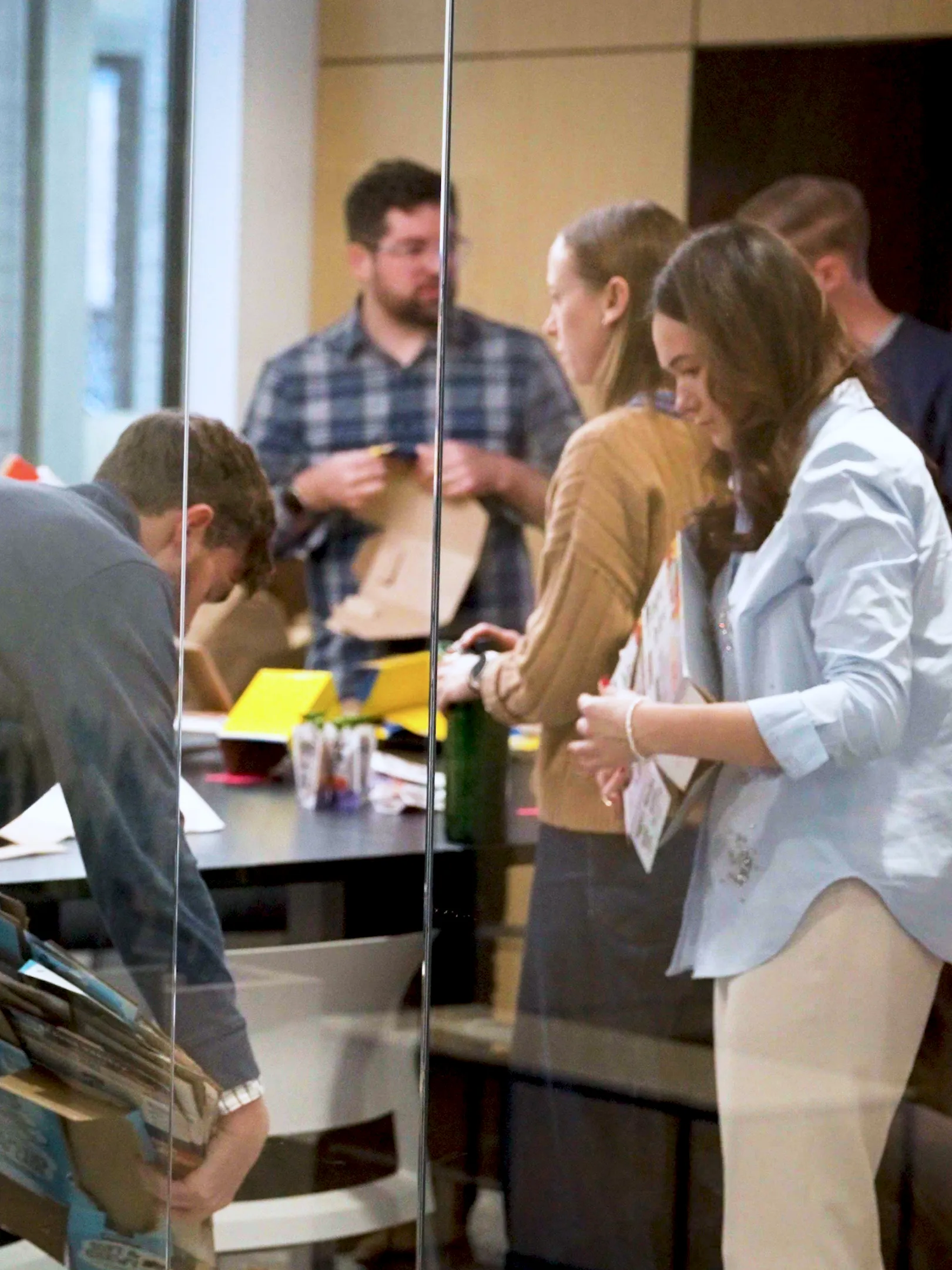 Several people stand and work around a table in an office, handling papers and folders. One person bends over picking up items while others are engaged in conversation or organizing materials.
