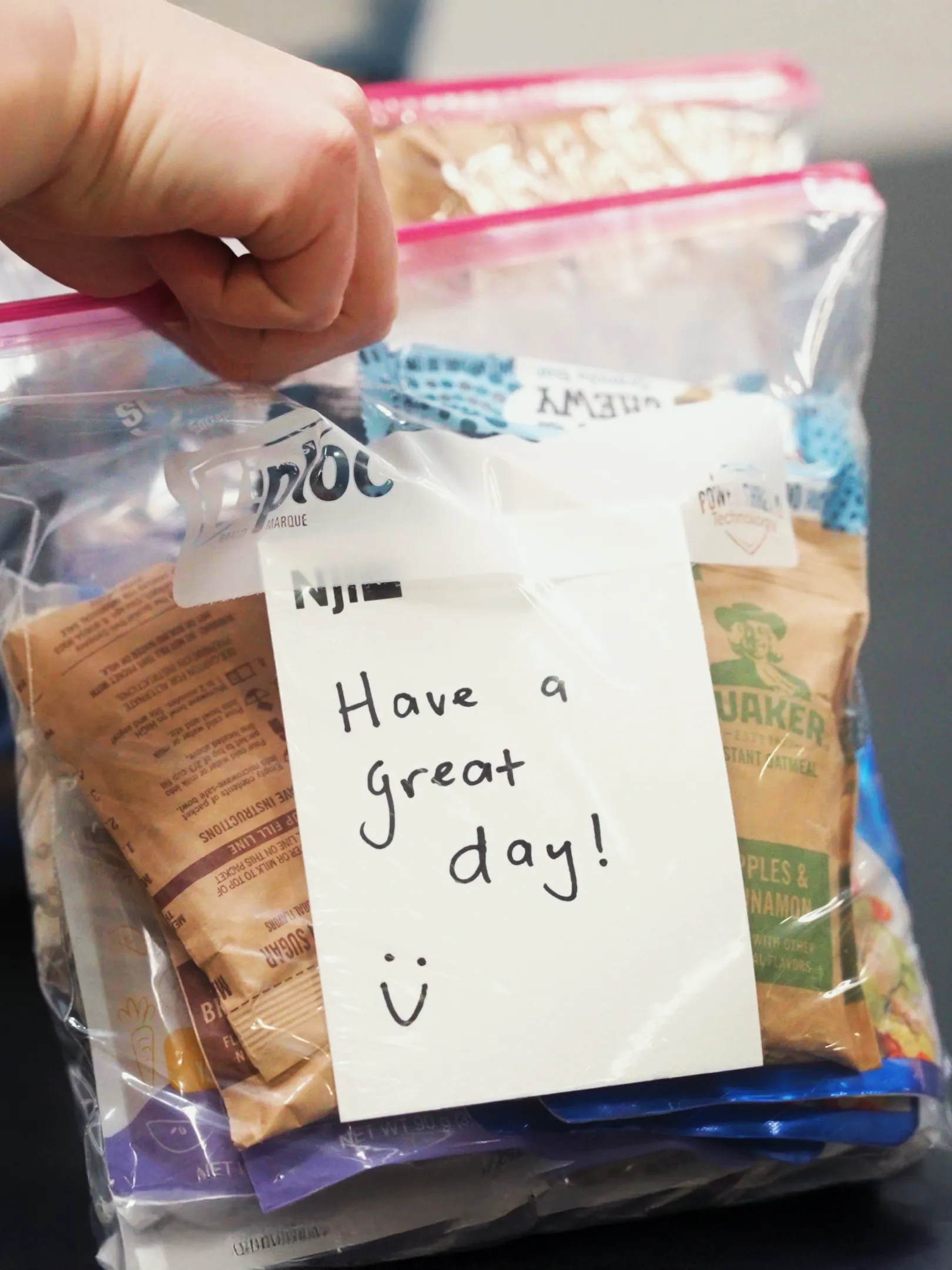 A hand holds a clear ziplock bag filled with snacks and oatmeal packets. A note inside reads, "Have a great day!" with a smiley face drawn at the bottom.