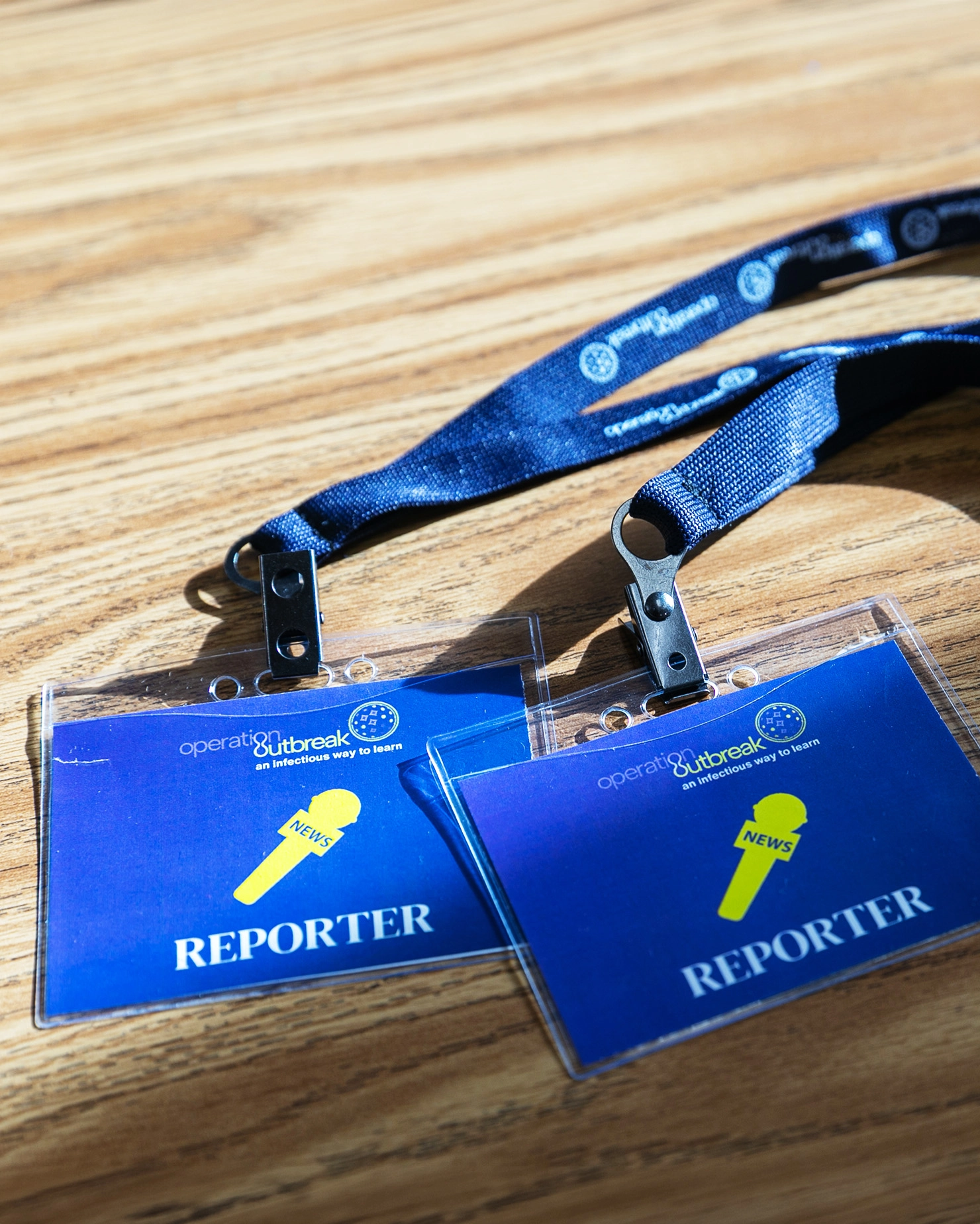 Two blue lanyards with "REPORTER" name badges, a yellow whistle, and the word "NEWS" lie on a wooden surface—ideal props for an experiential marketing or global awareness campaign led by PATH.