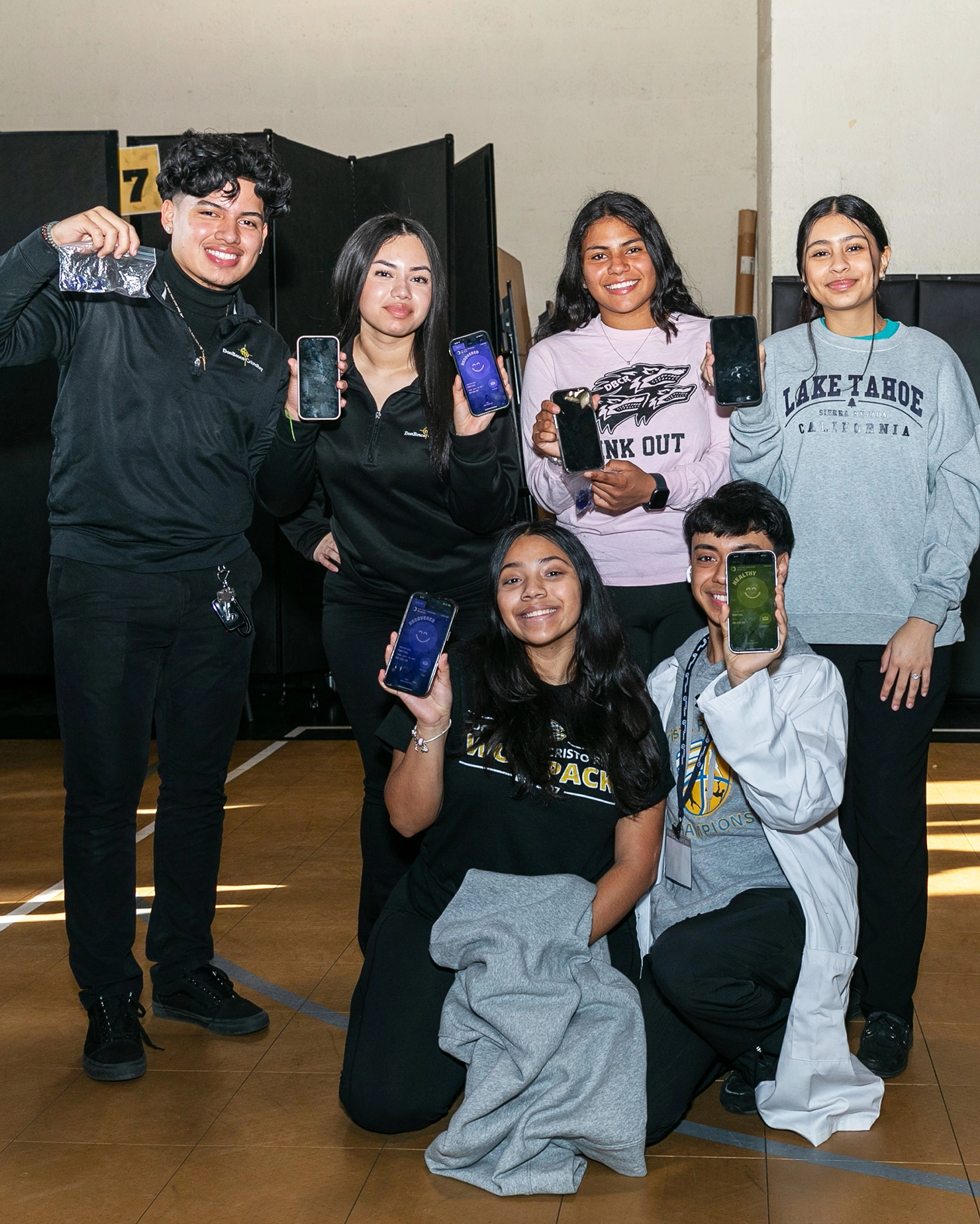 Six smiling teenagers pose indoors, holding up smartphones toward the camera for a global awareness campaign. Dressed casually, some wear school or sports logos as they kneel and stand on a gym floor, highlighting Health Advocacy against a dark backdrop.