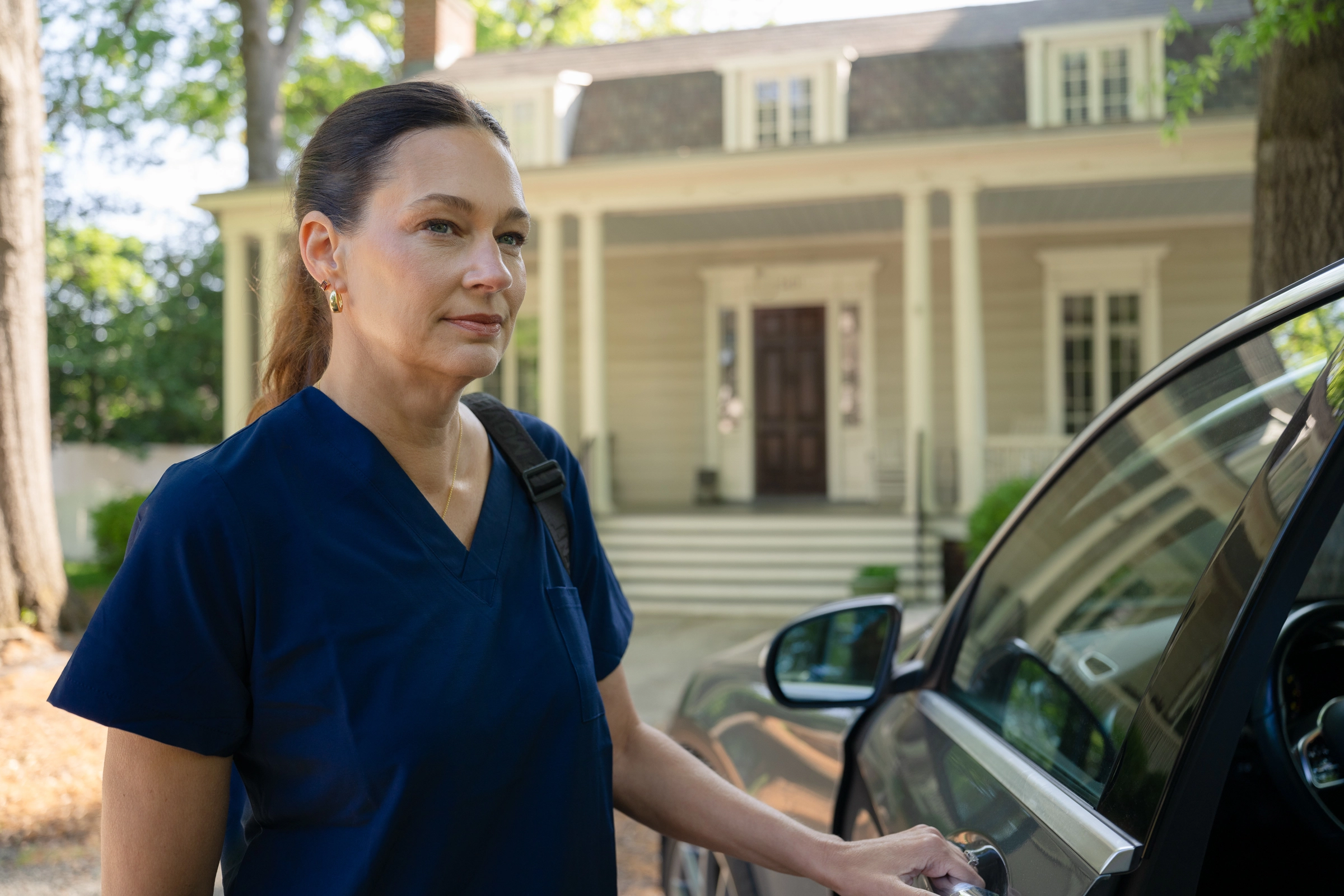 A woman in navy blue scrubs stands beside a car, holding the door handle, with a large house and trees in the background. She has a calm expression and a bag over her shoulder.