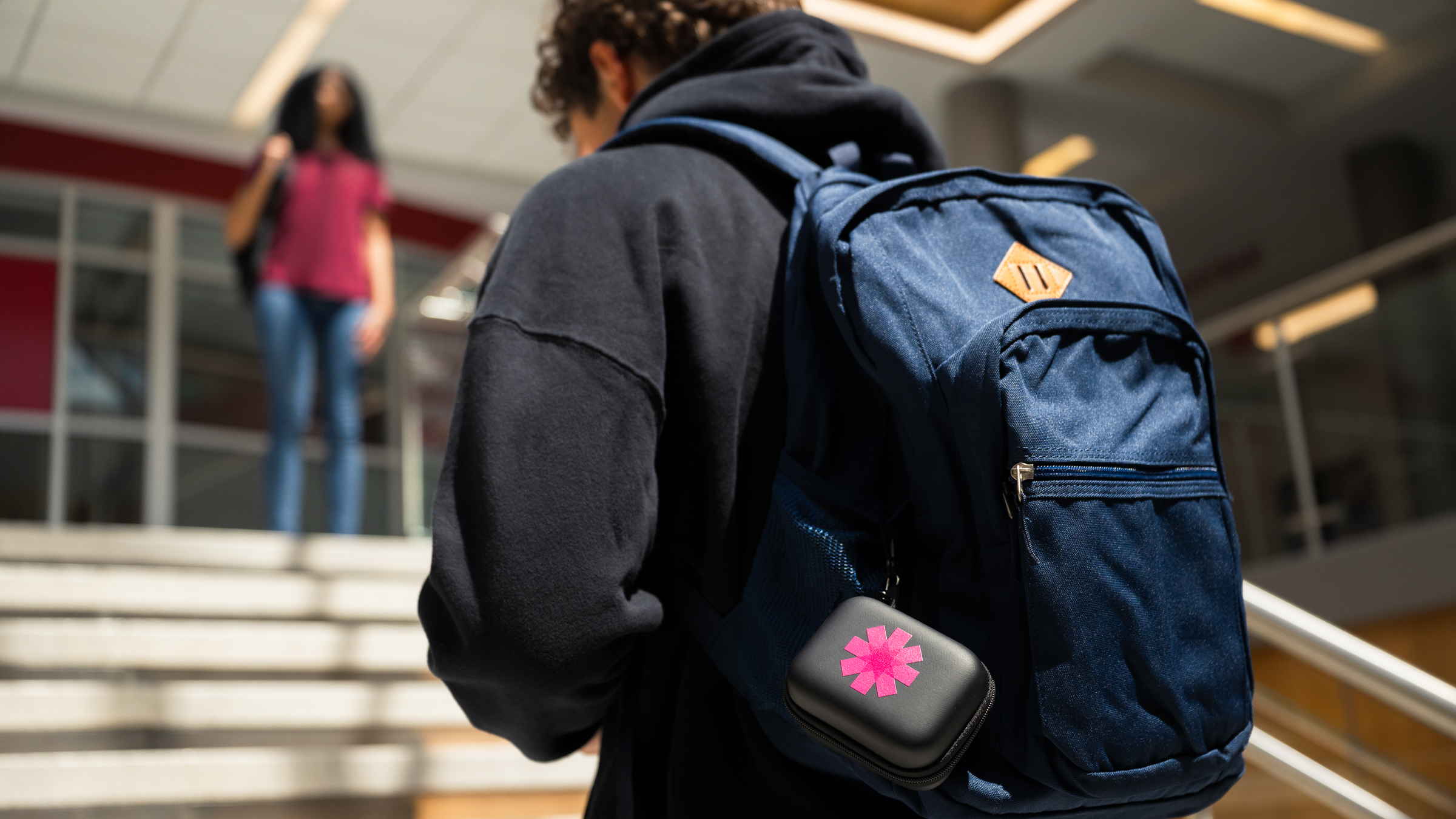 A person with curly hair and a navy blue backpack walks up indoor stairs. A small black case with a pink flower sticker is attached to the backpack. Another person stands at the top of the stairs in the background.