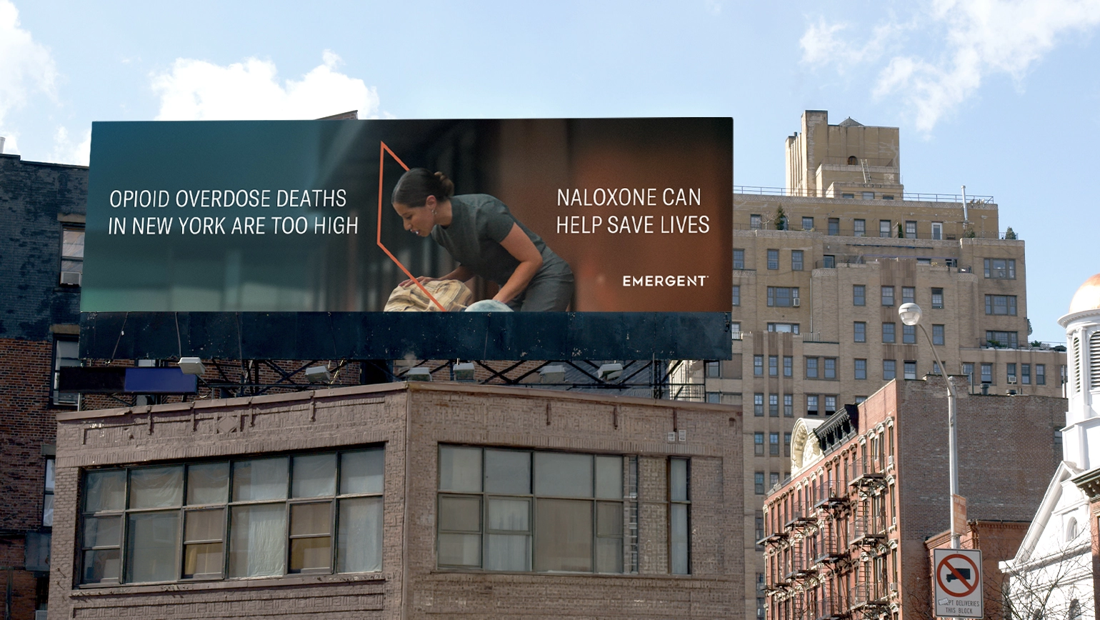 A billboard on a city building reads, “Opioid overdose deaths in New York are too high. Naloxone can help save lives.” The billboard shows a person assisting another person lying down. The Emergent logo is visible.