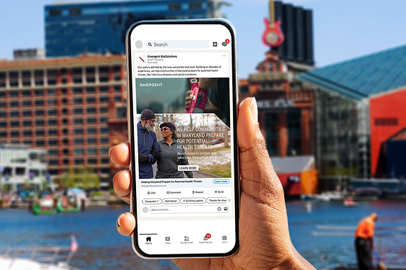 A hand holds a smartphone showing a community health webpage, with a city waterfront scene and buildings in the background. The phone screen features people and a message about health preparedness.