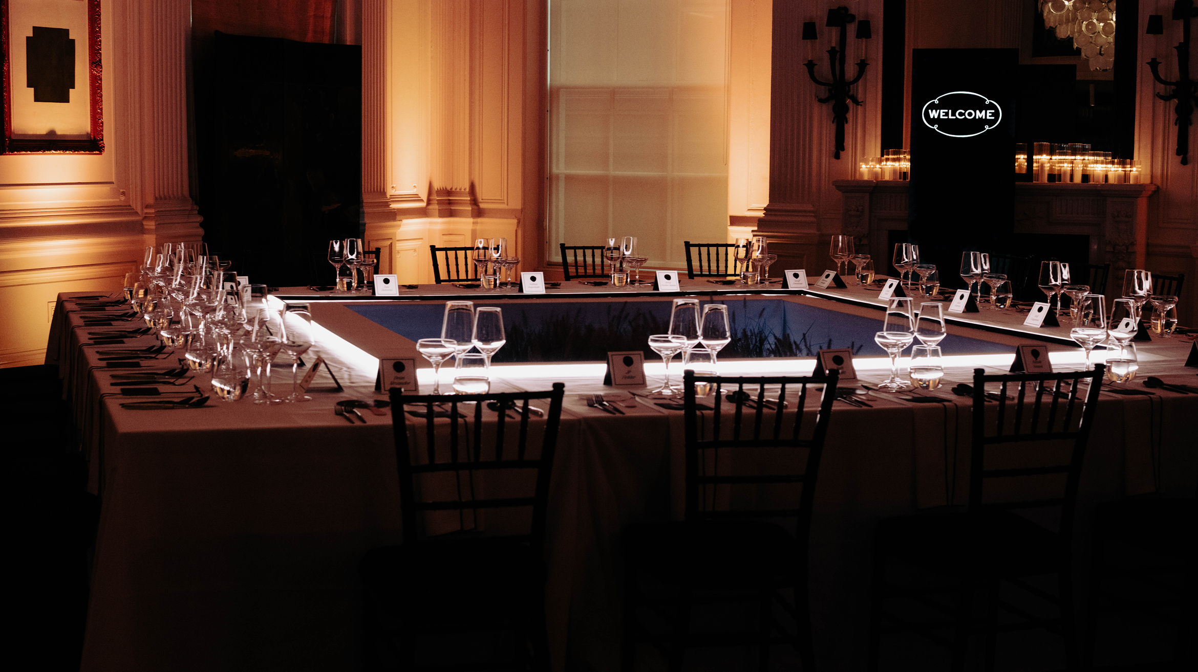 A square dinner table set for an event, surrounded by chairs, with place cards, wine glasses, and cutlery. A illuminated “WELCOME” sign is visible in the background. The room has warm, elegant lighting.