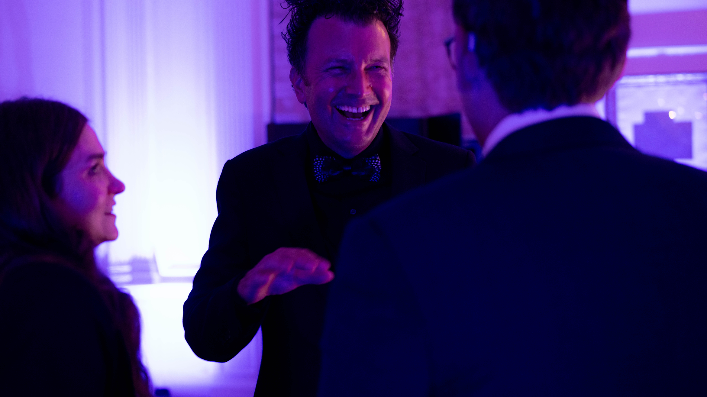A man in a suit and bow tie laughs and gestures while talking to two people at an indoor event with purple lighting.