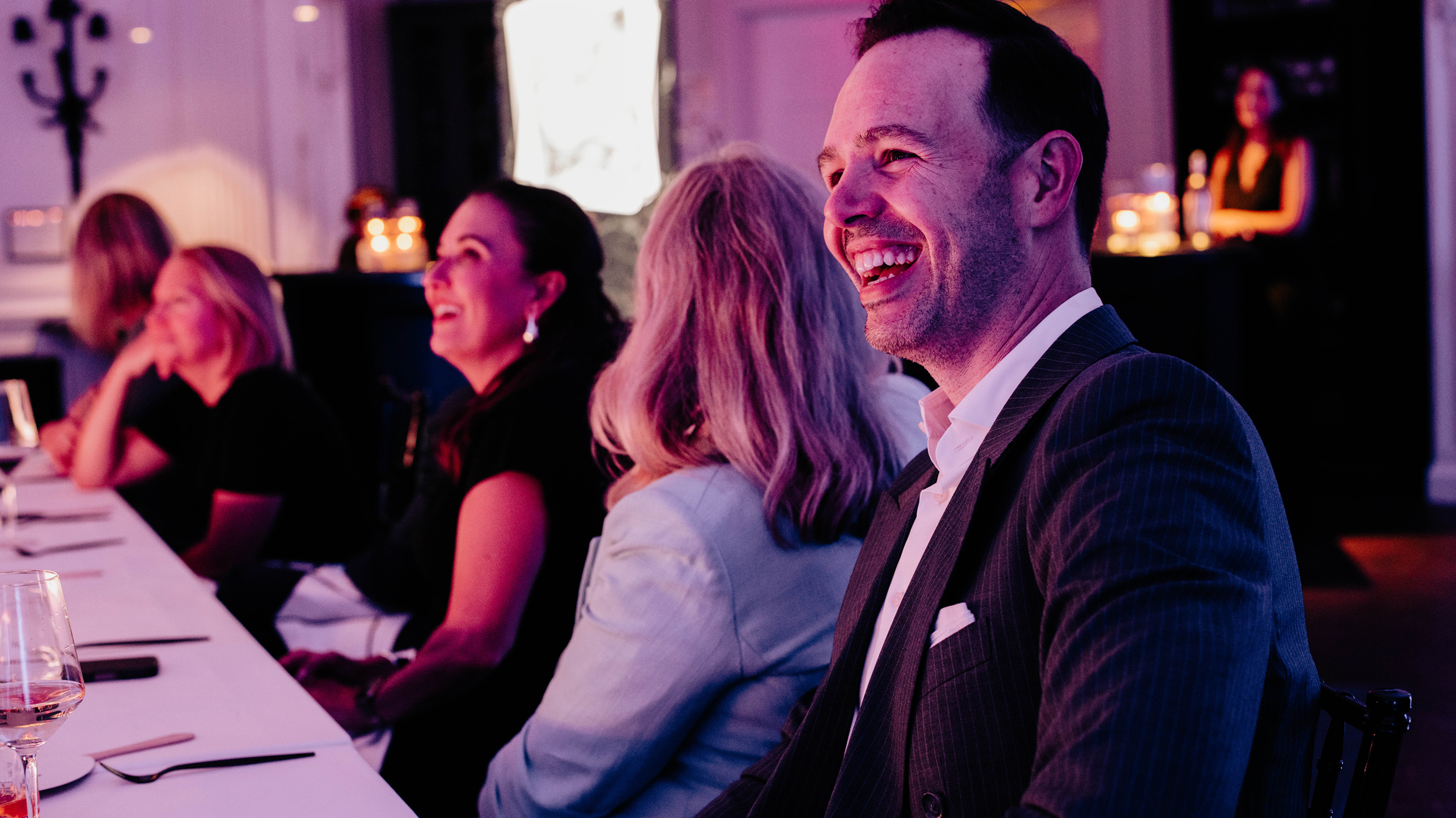 A group of people sit at a table, smiling and laughing in a warmly lit indoor setting, suggesting a joyful social or formal event.