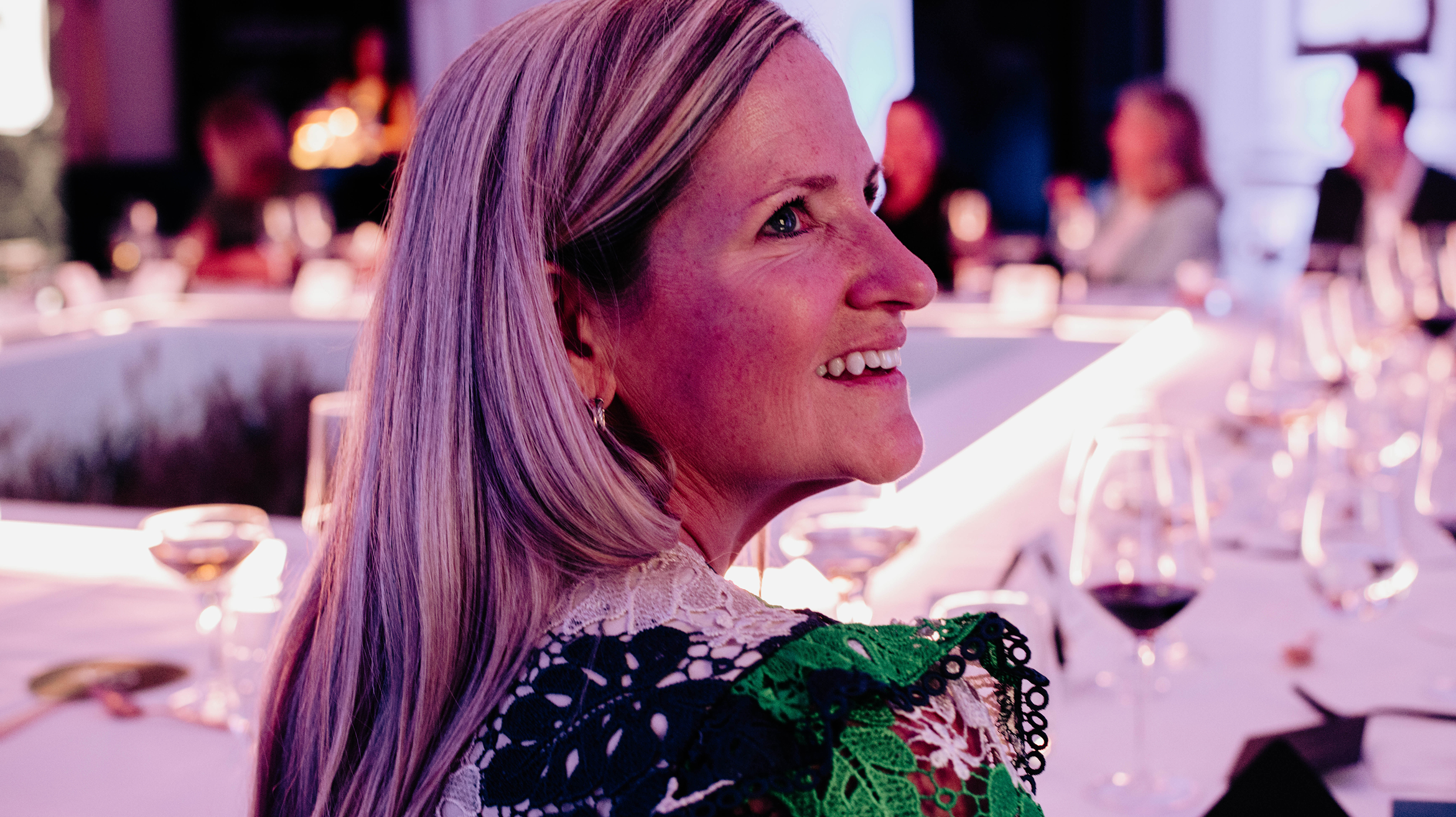 A woman with long blonde hair smiles while seated at a formal dinner table, surrounded by wine glasses and softly lit by purple and pink lighting. Other guests are blurred in the background.