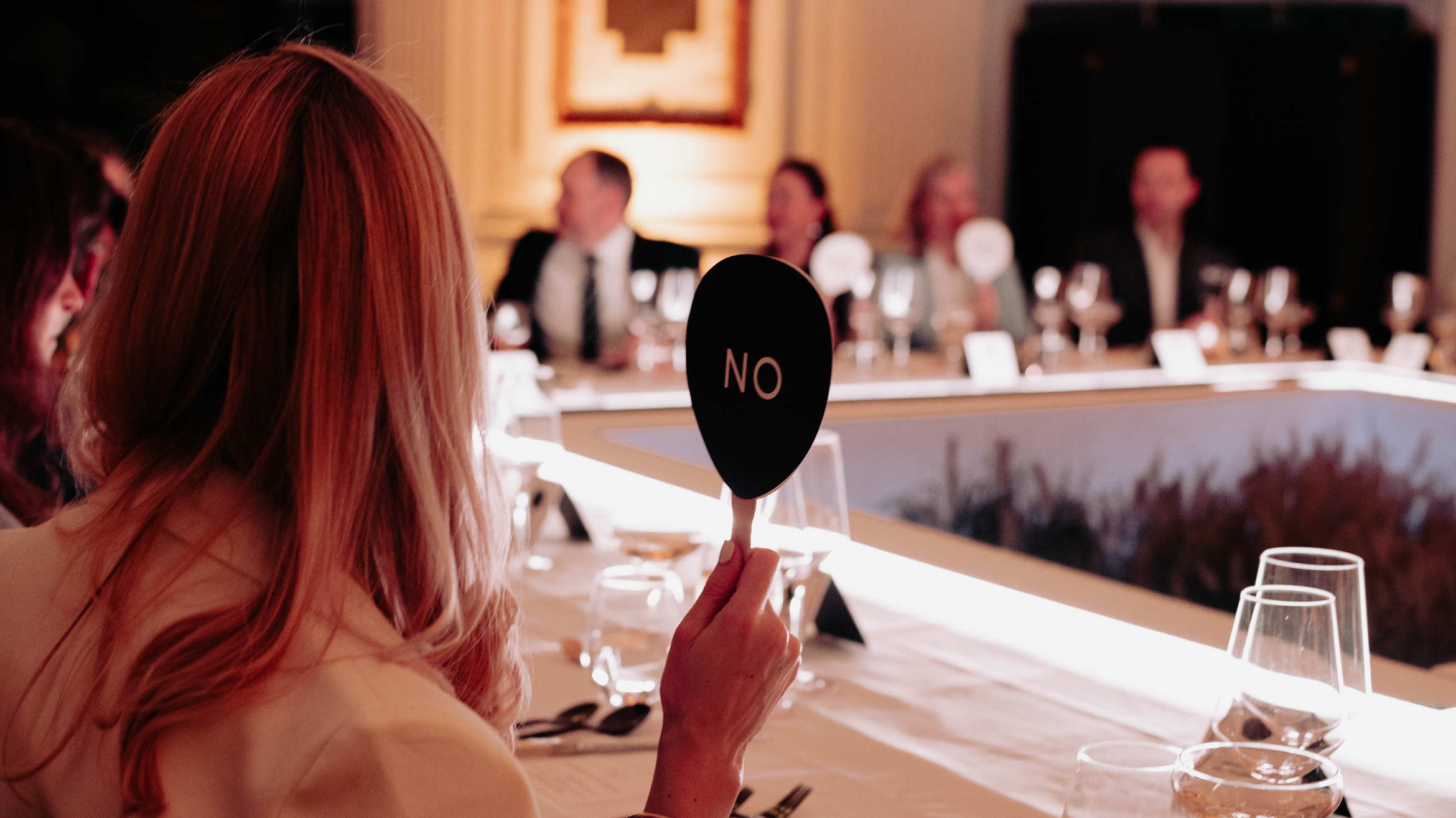 A person with long hair holds up a paddle that says “NO” at a formal dinner table, with several people seated in the background.