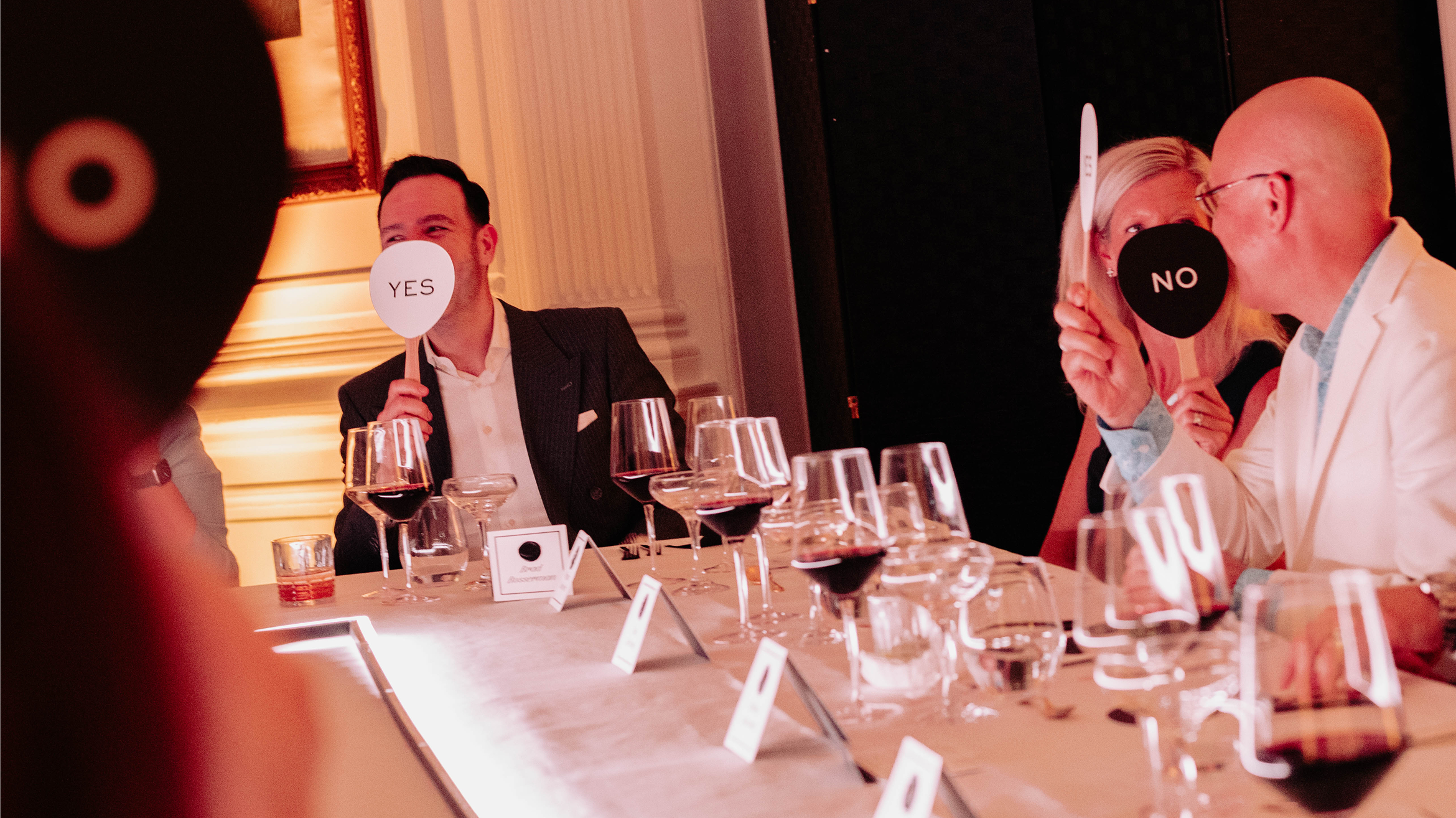 People at a formal dinner table hold up signs reading “YES” and “NO,” appearing to vote or make decisions, while smiling and socializing. There are wine glasses and table cards on the table.