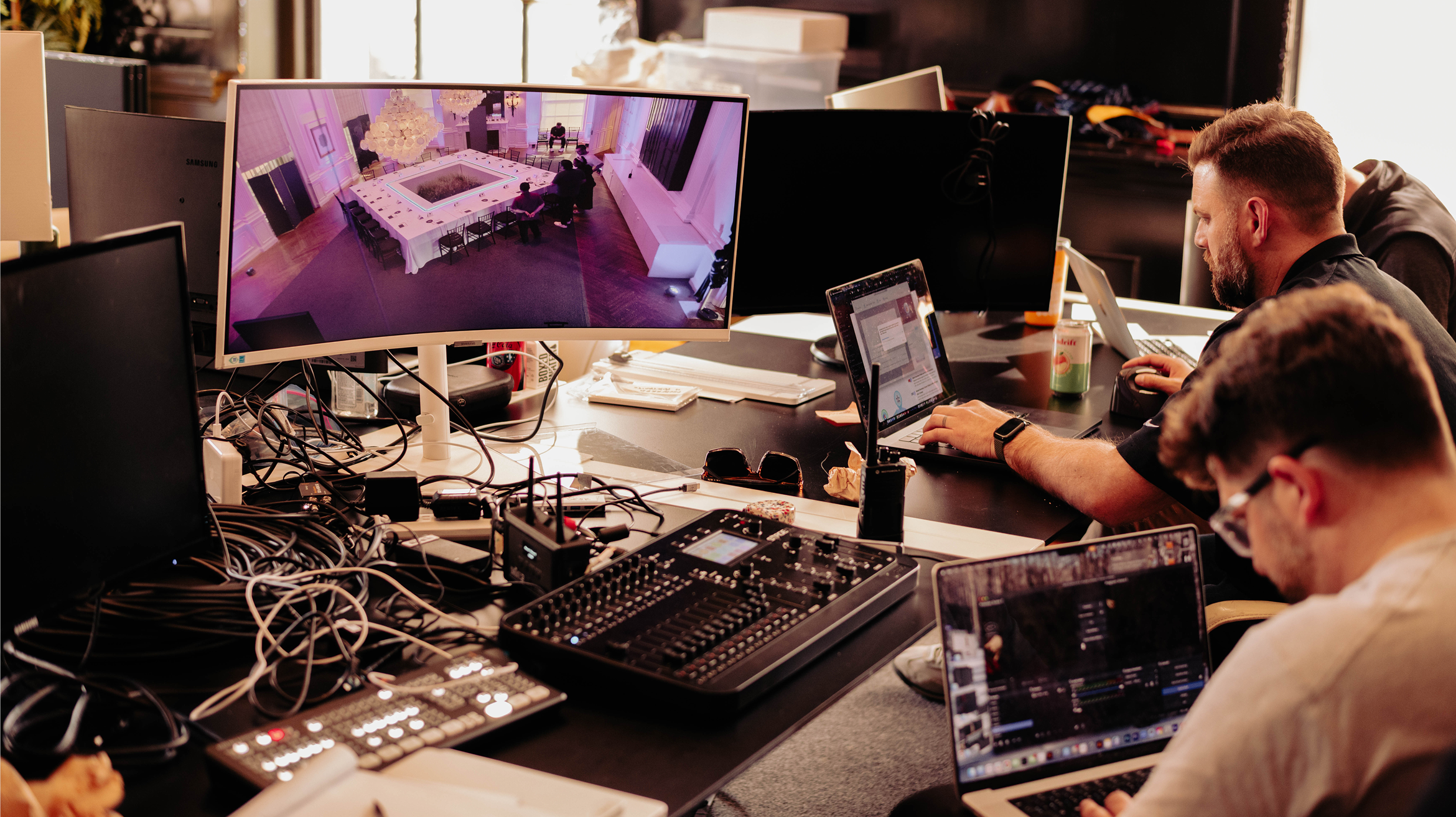Two people work at a desk with laptops and audio equipment. A large monitor displays a surveillance view of a conference room. Cables and electronics cover the workspace.