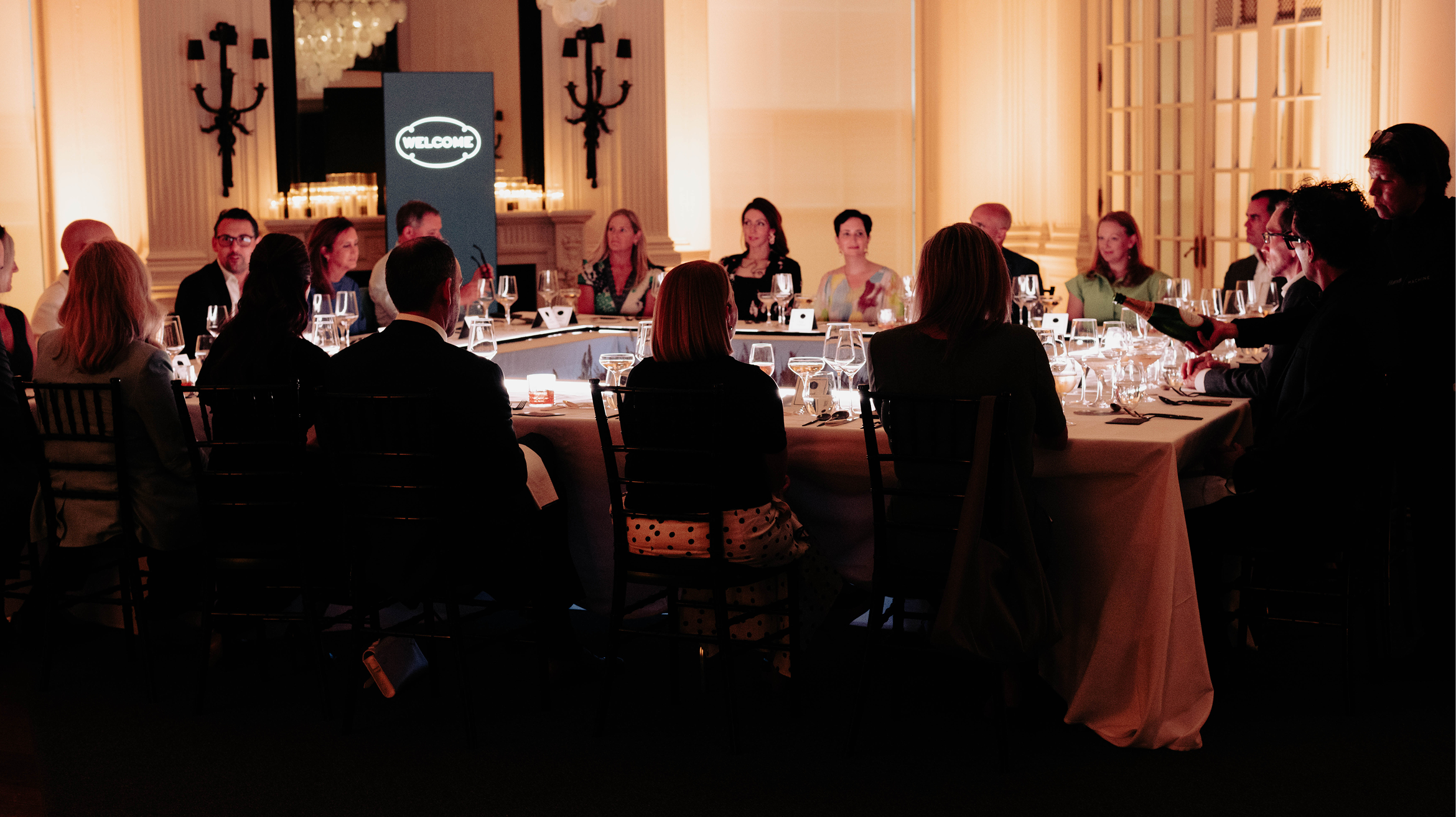 A group of people sit around a large, elegant dinner table in a warmly lit room. A screen in the background displays the word “WELCOME.” Glassware and place settings are arranged on the table.