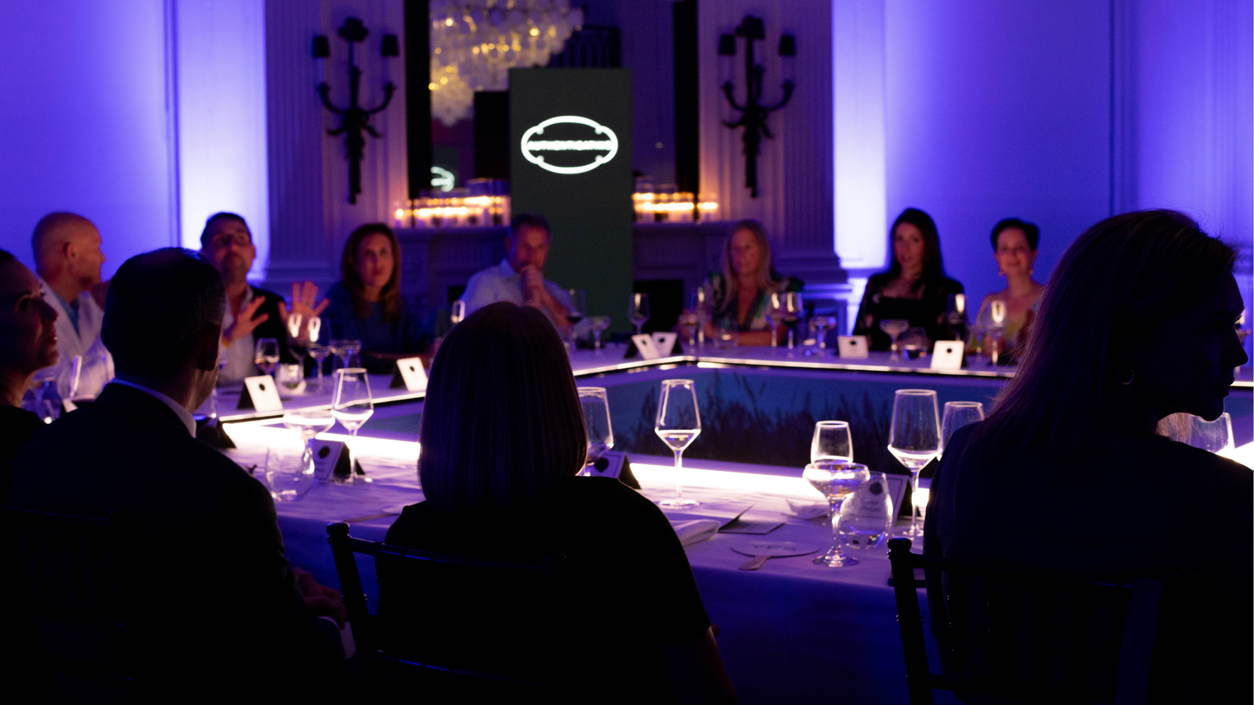 People sit around a U-shaped table set with glasses in a dimly lit room with blue lighting, engaged in conversation. A glowing light fixture and a backlit panel are visible in the background.