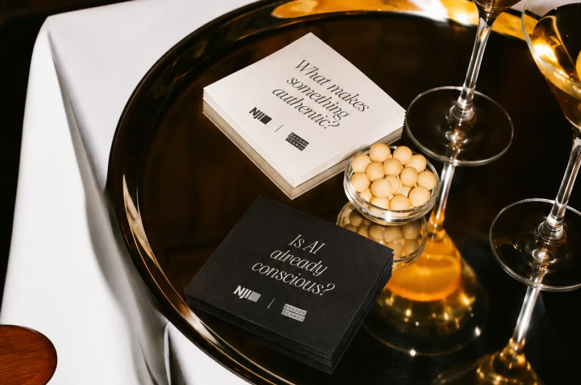 A gold tray holds wine glasses, a small bowl of round snacks, and two stacks of napkins with text: “What makes something authentic?” and “Is AI already conscious?”.