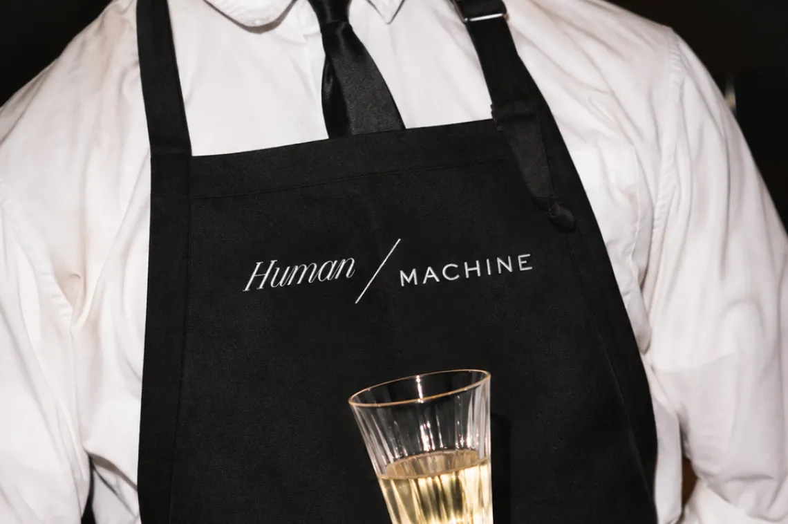 A person in a white shirt, black tie, and black apron with “Human / Machine” written on it holds a glass of white wine.