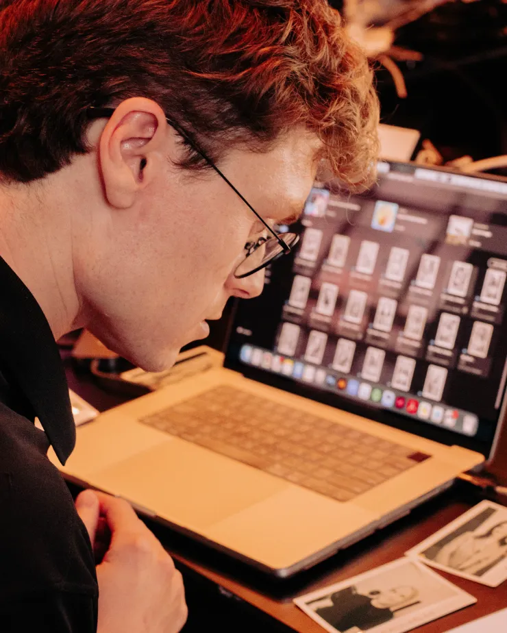 A person with glasses looks at printed photos on a desk, sitting in front of a laptop displaying image files.